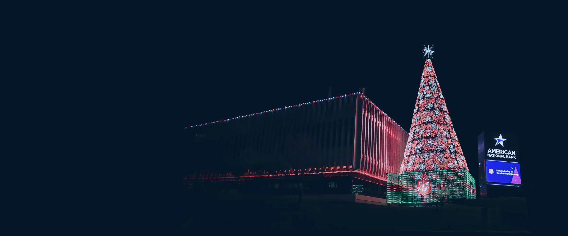 Photo of the American National Bank building with red holiday lights next to an LED Christmas tree and the American National Bank marquee.