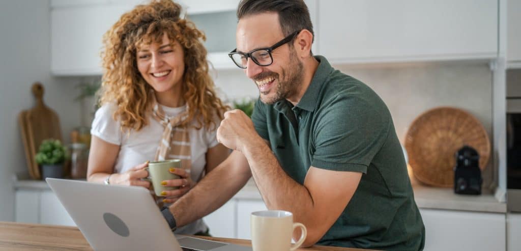 Happy couple sitting in kitchen drinking coffee and looking at their computer.