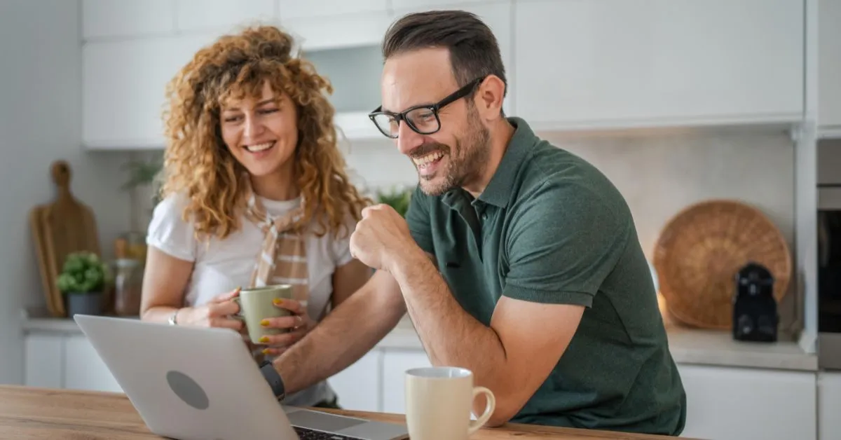 Happy couple sitting in kitchen drinking coffee and looking at their computer.