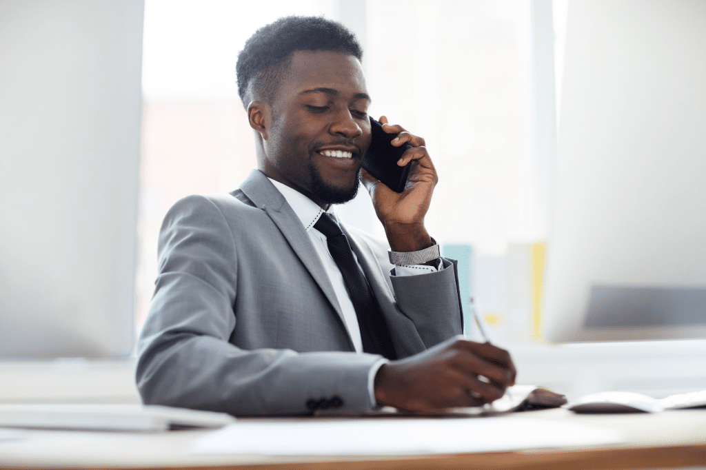 Happy young office manager talking by smartphone while sitting at desk and taking notes